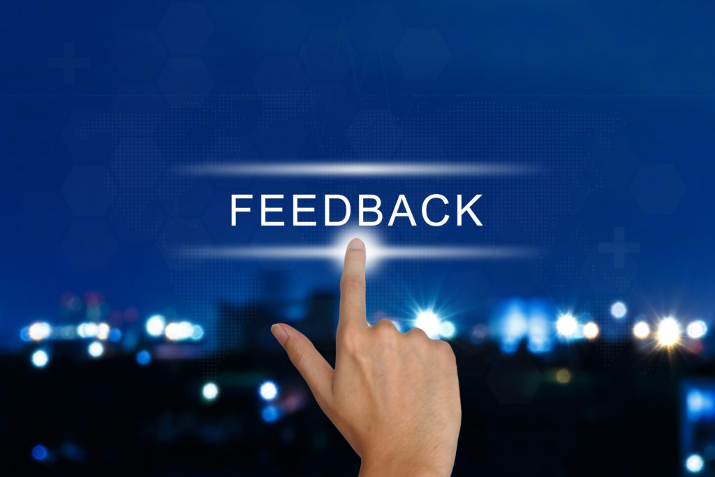 A hand points to the word "FEEDBACK" on a virtual interface, with a blurred cityscape and blue lights in the background.