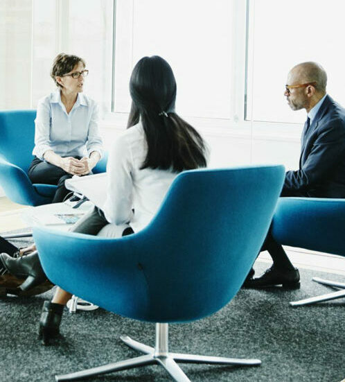 Three people sit in blue chairs having a conversation in a modern office with large windows and natural light.