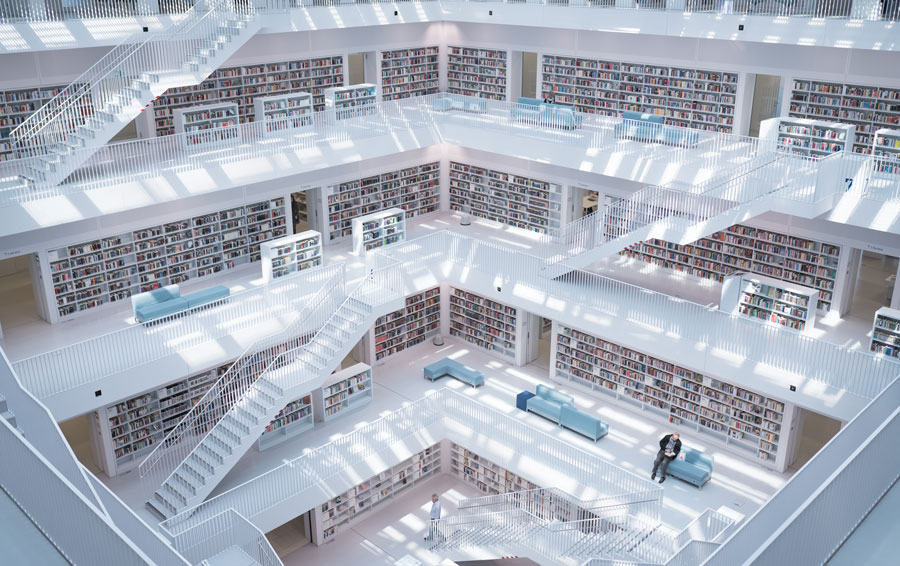 Modern multi-level library interior with white walls, open staircases, bookshelves lining the walls, and a few people walking and sitting on blue benches.