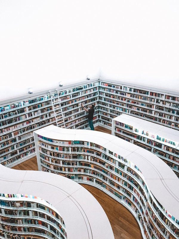 A person stands in a modern, white library surrounded by curved bookshelves filled with books, reaching for a book on an upper shelf.