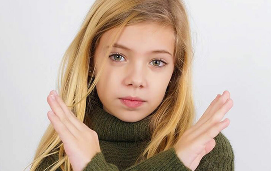 A young girl with long blonde hair wearing a green sweater holds her arms in an X shape in front of her chest, indicating refusal or disagreement.