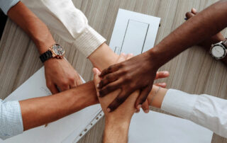 Five people seated at a table stack their hands in the center over documents, suggesting teamwork or collaboration.