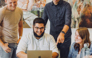 Four people gathered around a laptop, smiling and looking at the screen, with abstract artwork on the wall behind them.