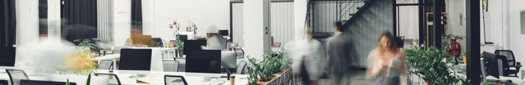 Modern open-plan office with white desks, computers, green plants, and blurred motion of people walking. The space features high ceilings, large windows, and industrial-style decor.