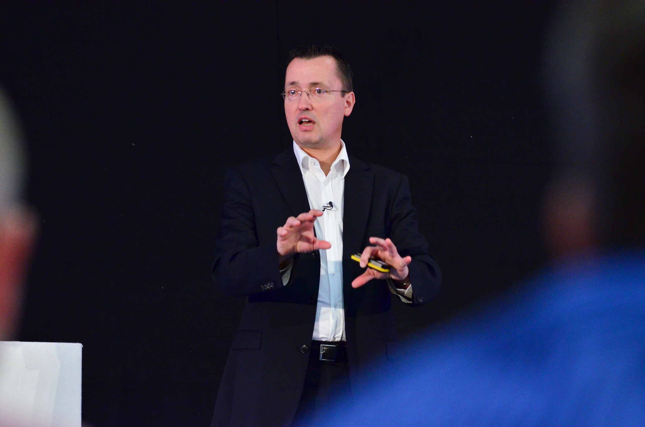 Stefan Osthaus, dressed in a suit, stands and gestures while speaking on stage against a black background, holding a small yellow object in one hand.