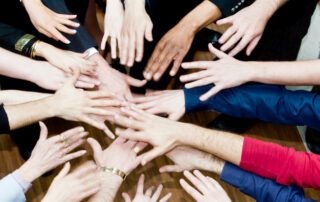 A group of people stand in a circle and reach their hands toward the center, overlapping above a wooden floor.